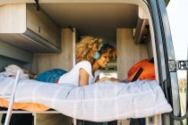 Mulher afro-americana alegre sorrindo e ouvindo música em fones de ouvido enquanto estava deitada na cama na caravana e navegando nas mídias sociais no laptop — Fotografia de Stock