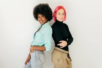 Cheerful young pink haired woman and African American girlfriend in stylish outfit standing together back to back on white background — Stock Photo