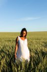 Sonriente joven dama negra en vestido de verano blanco paseando por el campo de trigo verde mientras mira a la cámara durante el día bajo el cielo azul - foto de stock