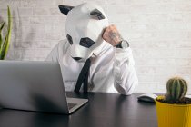 Анонімний втомлений чоловічий підприємець у масці ведмедя панди та біла сорочка, що працює за столом з нетбуком на робочому місці — стокове фото