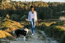 Full body of happy ethnic woman with Border Collie dog walking together on trail among grassy hills in sunny spring evening — Stock Photo