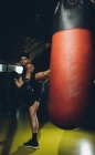 Jeune homme asiatique concentré entraînement kick boxing exécution coups de pied de grève tout en exerçant avec un sac de boxe lourd dans une salle de gym moderne — Photo de stock