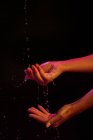 Schnittansicht einer anonymen Frau, die sich unter Neonlicht vor schwarzem Hintergrund die Hände mit Spritzwasser wäscht — Stockfoto