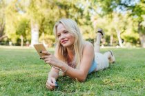 Sorrindo encantadora fêmea deitada na grama no parque tirando selfie no smartphone e ouvindo música em fones de ouvido no verão — Fotografia de Stock