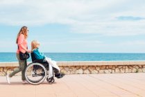 Seitenansicht einer erwachsenen Tochter, die im Sommer mit einer älteren Mutter im Rollstuhl entlang der Strandpromenade am Meer spaziert — Stockfoto
