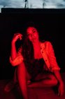 Sinnliche junge hispanische ethnische Frau in Dessous blickt in die Kamera, während sie nachts auf der Terrasse unter rotem Neonlicht ruht — Stockfoto