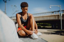 Joven fitness mujer afroamericana descansando y atando los cordones después de hacer ejercicio en el banco del parque - foto de stock