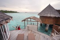 Сверху вид на спину неузнаваемой женщины в красном платье с поднятыми руками, танцующей у океана с безликим партнером в повседневной одежде, расслабляющей на Мальдивах — стоковое фото