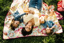 Вид сверху счастливой молодой женщины и восхитительных маленьких сестер в похожих платьях, лежащих на одеяле на зеленой траве, проводя летний день вместе в парке — стоковое фото