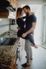Homem barbudo alegre abraçando mulher sorridente enquanto se inclina no armário com pia na cozinha acolhedora em casa — Fotografia de Stock