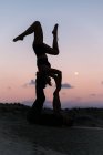Vista lateral de una mujer flexible balanceándose boca abajo mientras practica acroyoga con su pareja masculina contra el cielo al atardecer en las montañas - foto de stock