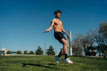 Male athlete in naked torso jumping with skipping rope and looking away on walkway during cardio training in park — Stock Photo