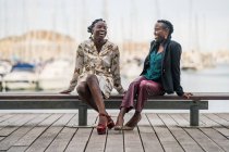 Стильна мелодія, що посміхається афроамериканцям, проводить час разом, сидячи на дерев'яній низькій лавці в парку в яскравий день. — стокове фото