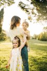 Menina pré-escolar bonito sorrindo e olhando para a câmera enquanto abraçando a mãe com a irmãzinha a mãos durante o dia de verão juntos no parque verde — Fotografia de Stock