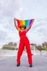 From below of stylish African American female in trendy wear raising flag with rainbow ornament while looking up on roadway — Stock Photo