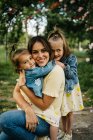 Felice giovane madre con simpatiche figlie in vestiti simili godendo soleggiata giornata estiva insieme mentre seduti nel parco — Foto stock
