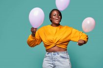 Excited African American female in jeans and yellow top standing near falling balloons against blue background in studio — Stock Photo