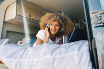 Mulher afro-americana alegre sorrindo e ouvindo música em fones de ouvido enquanto estava deitada na cama na caravana e navegando nas mídias sociais no laptop — Fotografia de Stock