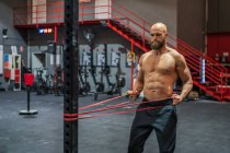 Hombre barbudo muscular tirando de la cuerda elástica con esfuerzo durante el entrenamiento funcional en el gimnasio moderno - foto de stock