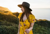 Приваблива молода жінка в жовтому сараї і капелюсі стоїть на травоїдному лузі в сонячній сільській місцевості — стокове фото
