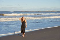 Full body smiling female in summer dress standing on sandy seashore and looking away — Stock Photo