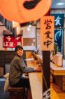 Ásia mulher no casual desgaste sentado no madeira contador enquanto espera para ordem no ramen bar — Fotografia de Stock