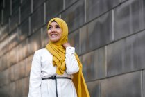 Низкий угол модной мусульманки в желтом хиджабе, стоящей на улице и смотрящей в сторону — стоковое фото