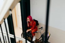 From above of young stylish female in red suit with backpack talking on smartphone while standing with bicycle on staircase — Stock Photo