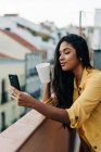 Junge hispanische Frau genießt frischen Kaffee und benutzt Mobiltelefon, während sie sich abends auf dem Balkon entspannen — Stockfoto