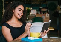 Smiling young curly haired Hispanic female flipping notebook while resting at table with cup of coffee on cafe terrace — Stock Photo