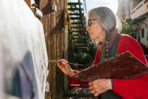 De dessous vue latérale des artistes féminines âgées en peinture de masque de protection sur toile dans la cour arrière par jour ensoleillé en été — Photo de stock