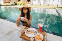 Cheerful female tourist leaning on poolside while drinking coffee against tray with yummy breakfast in sunlight — Stock Photo