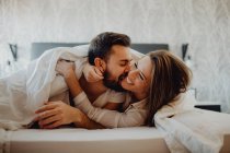 Cheerful young man and woman smiling and cuddling while lying on comfortable bed at home together — Stock Photo