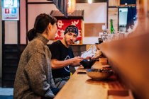 Vista lateral da mulher e do homem se comunicando enquanto come comida asiática no balcão de madeira no café moderno — Fotografia de Stock