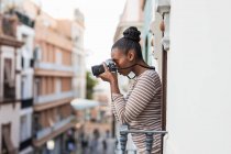 Вид збоку етнічна жінка в одязі зі смугастим орнаментом з професійним фото пристроєм, що дивиться на балкон вдень — стокове фото