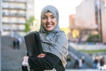 Femme entrepreneur musulmane joyeuse dans le hijab et avec le dossier debout regardant loin dans la rue — Photo de stock