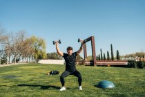 Sportsman in sportswear working out with dumbbells while performing forward lunge and breathing on meadow in city — Stock Photo