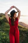 Attractive young female in red sundress and hat standing with arms raised on verdant grassy meadow in sunny countryside — Stock Photo