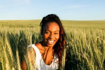 Jeune dame noire en robe d'été blanche se promenant sur le champ de blé vert tout en regardant la caméra de jour sous le ciel bleu — Photo de stock