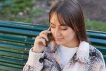 From above modern millennial female in stylish spring outfit sitting on bench and answering phone call while resting on urban street looking away — Stock Photo