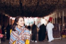Jeune femme orientale en costume traditionnel et foulard souriant et regardant loin tout en se tenant debout sur un fond flou du vieux bazar souk Manama dans la ville de Manama au Bahreïn — Photo de stock