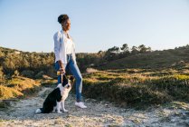 Vista laterale del corpo pieno di donna etnica felice con il cane Border Collie che cammina insieme sul sentiero tra le colline erbose nella soleggiata serata primaverile — Foto stock