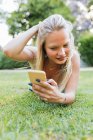 Улыбающаяся очаровательная женщина, лежащая на траве в парке и слушающая музыку в наушниках летом — стоковое фото