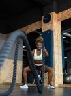 Fuerte atleta afroamericana en ropa deportiva haciendo ejercicio con cuerdas de batalla mientras mira hacia adelante durante el entrenamiento de alta intensidad en el gimnasio - foto de stock