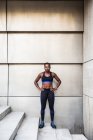 Mujer afroamericana positiva en ropa deportiva que mantiene las manos en la cintura y mira la cámara mientras está de pie en las escaleras durante el entrenamiento al aire libre - foto de stock