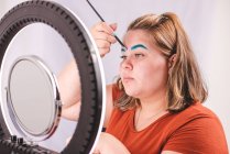 Femme en surpoids avec palette appliquant des pigments colorés sur le visage tout en regardant le miroir près de la lumière annulaire en studio — Photo de stock