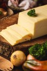 Bloque de queso en soporte de madera cerca de cebollas crudas y bolsa tejida contra espátulas orgánicas y hojas de albahaca - foto de stock