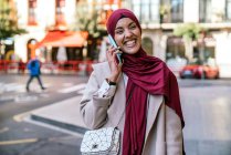 Mujer musulmana alegre en hijab y con bolsas de compras caminando por la calle y hablando en un teléfono inteligente - foto de stock