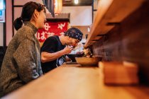 Seitenansicht einer Frau und eines Mannes, die beim Essen asiatischer Speisen an der Holztheke im modernen Café miteinander kommunizieren — Stockfoto