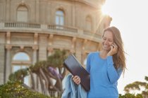 Heureuse femme adulte en tenue décontractée parlant sur smartphone avec dossier à la main tout en se tenant près de plantes vertes et vieux bâtiment dans le parc par temps ensoleillé — Photo de stock